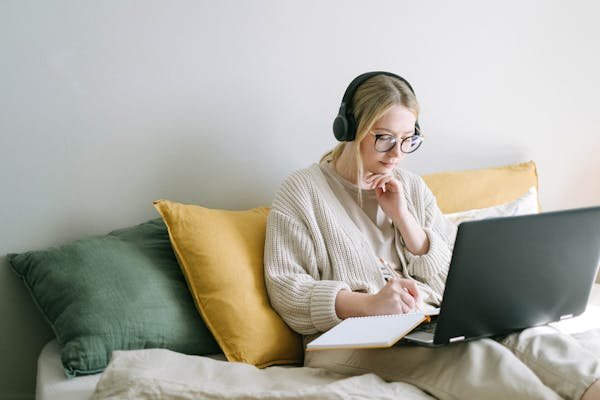 A focused woman in glasses and headphones works on a laptop from a cozy bed.