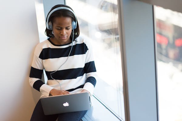 African American woman wearing headphones using laptop by a sunny window indoors.