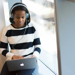 African American woman wearing headphones using laptop by a sunny window indoors.