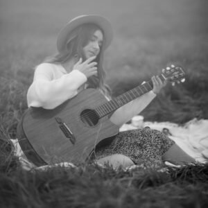 Black and white photo of a woman playing guitar in a grass field, wearing a hat.