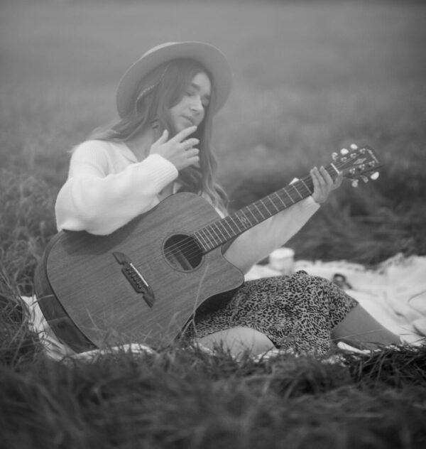 Black and white photo of a woman playing guitar in a grass field, wearing a hat.