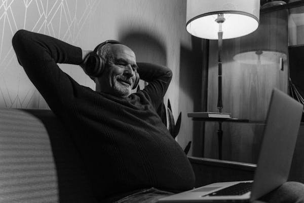 Elderly man enjoying music with headphones, sitting comfortably with a laptop in grayscale image.