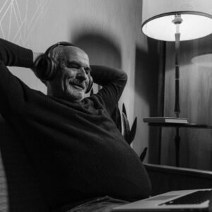 Elderly man enjoying music with headphones, sitting comfortably with a laptop in grayscale image.