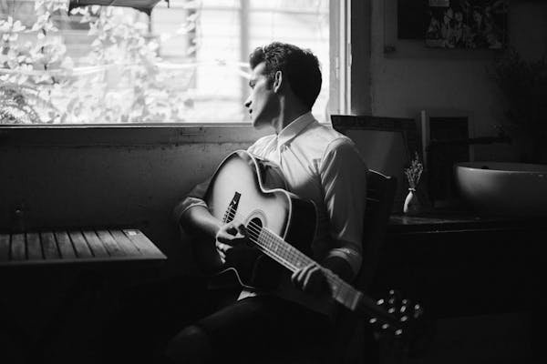 Thoughtful man with guitar looking out window in black and white.