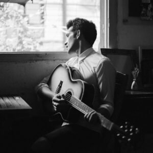 Thoughtful man with guitar looking out window in black and white.